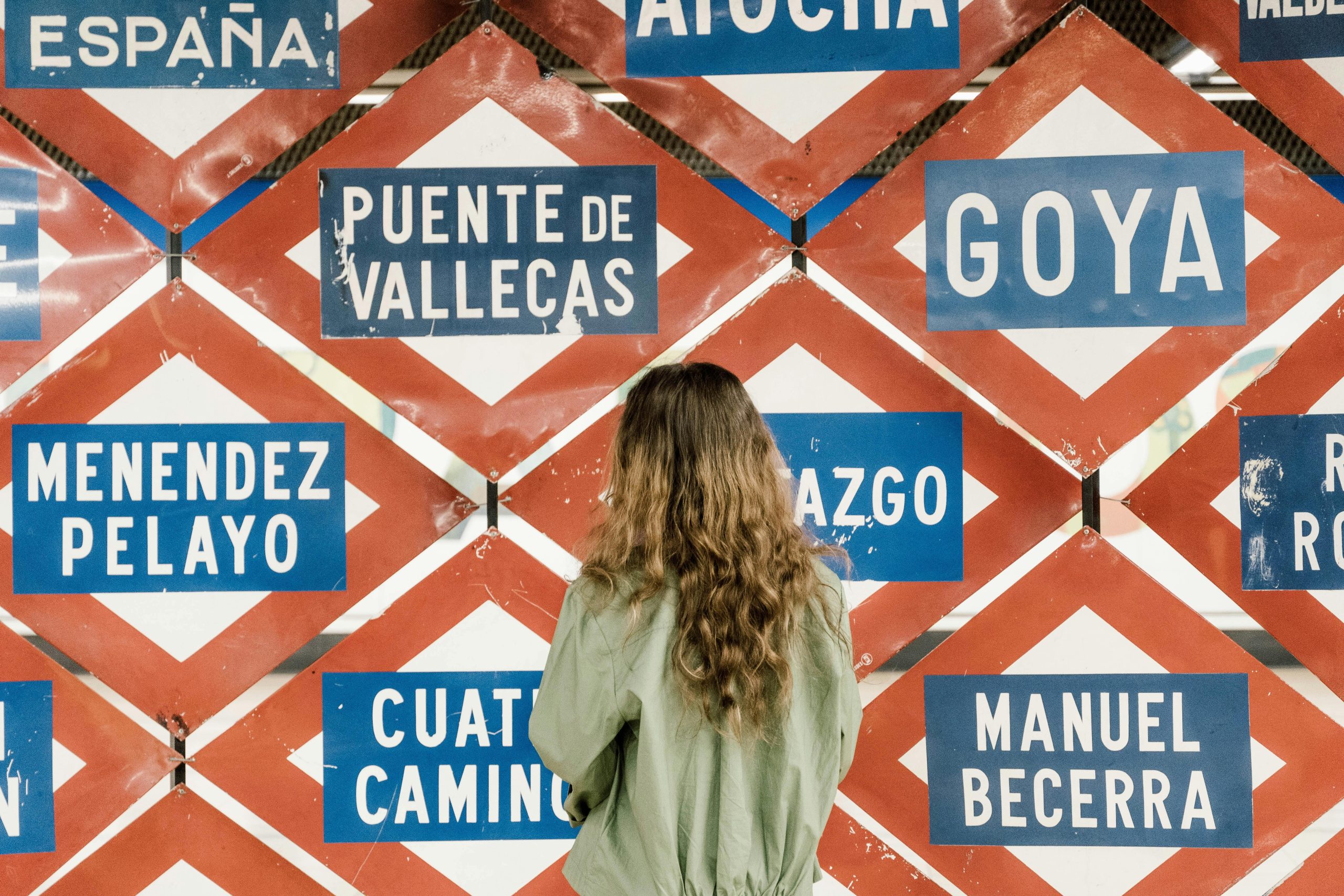 Mujer de espaldas mirando un cartel con varias estaciones del metro de Madrid. Un instante de pausa en la ciudad que nunca se detiene, donde cada parada esconde una historia por descubrir.